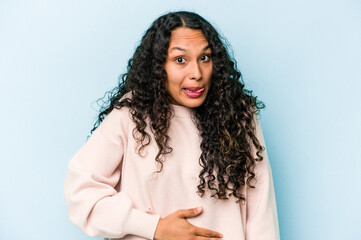 Young hispanic woman isolated on blue background touches tummy, smiles gently, eating and satisfaction concept.