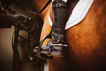 Fotobehang The chestnut horse is wearing horse ammunition - a stirrup, a brown old saddle, a white saddlecloth, a bridle, and a rider in black boots is sitting in the saddle. Horse riding. Equestrian sports. ©  Valeri Vatel