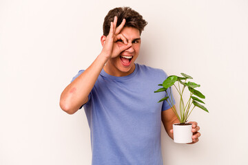 Young caucasian man holding a plant isolated on white background excited keeping ok gesture on eye.