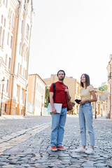 Obraz premium Happy woman holding binoculars near boyfriend with map and backpacks on city street.