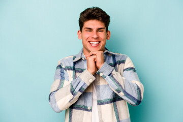 Young caucasian man isolated on blue background keeps hands under chin, is looking happily aside.