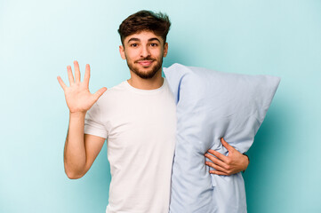 Young hispanic man wearing a pajama holding pillow isolated on blue background smiling cheerful showing number five with fingers.