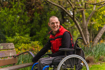 Portrait of a person with a disability in a red vest in a public park in the city. Sitting in the wheelchair next to a park chair