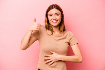 Young caucasian woman isolated on pink background touches tummy, smiles gently, eating and satisfaction concept.