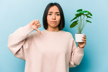 Young hispanic woman holding a plant isolated on blue background showing a dislike gesture, thumbs...