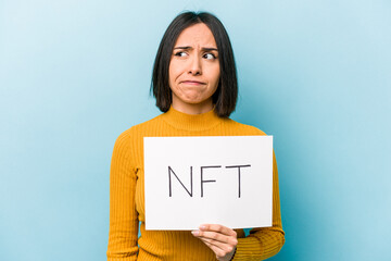 Young hispanic woman holding NFT placard isolated on blue background confused, feels doubtful and unsure.