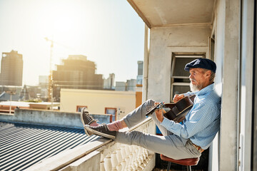 His daily ritual of playing guitar. Shot of a peaceful senior man sitting on his balcony while playing acoustic guitar with the city in the background.