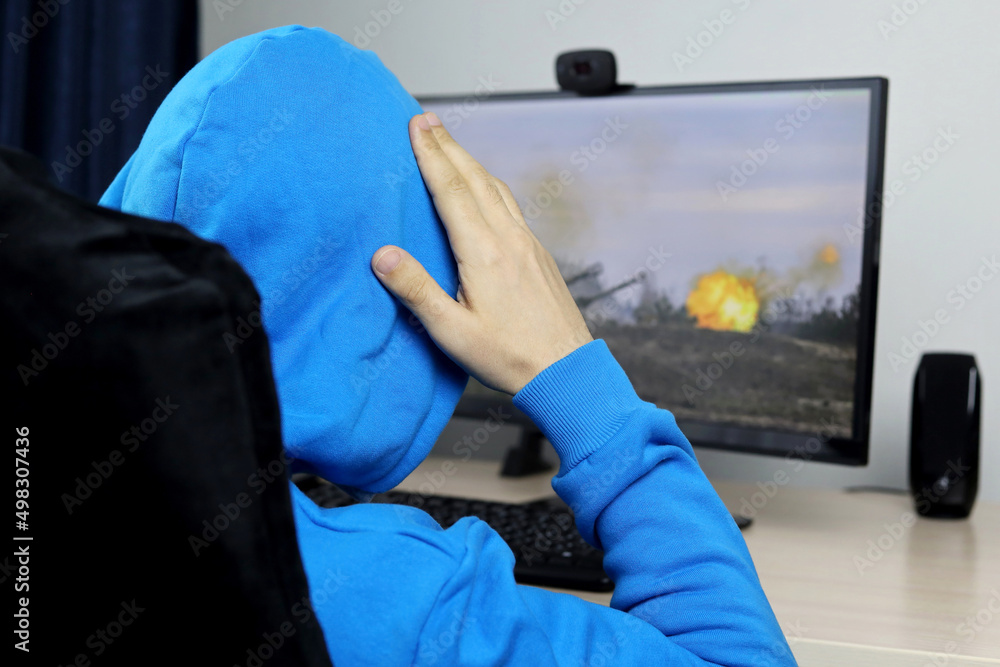 Wall mural Man in hoodie sitting at desktop PC and looking at the firing artillery. Video news about the war in Ukraine - Wall murals