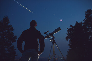 Silhouette of a man, telescope, stars, planets and shooting star under the night sky.