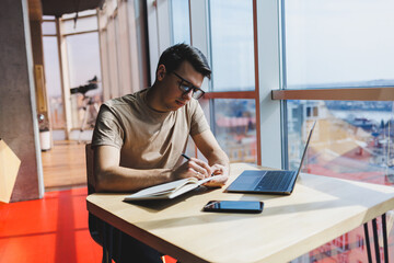 A European businessman sits with a laptop during lunch in a cafe. The concept of remote and freelance work. Smiling adult successful man in glasses is sitting at a wooden table. sunny day