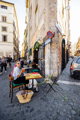 Woman sitting at traditional restaurant on the beautiful cozy street in Rome. Concept of Italian gastronomy and travel. wide street view