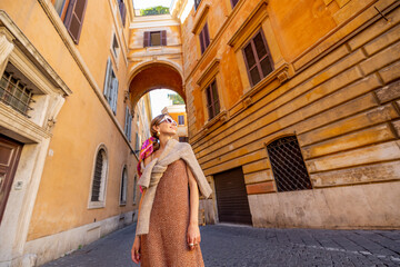 Woman walks on narrow old street with beautiful buildings and arch ahead in Rome. Portrait of a happy stylish woman in Rome. Wide angle view