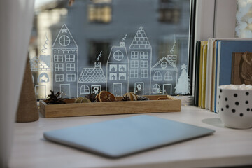 Laptop and hot drink on white wooden table near window with beautiful drawing at home. Christmas decor
