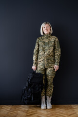 full length of woman in military uniform standing with black backpack near dark wall.