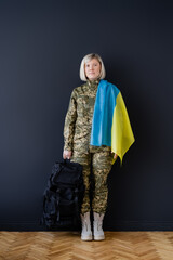 full length of military woman with ukrainian flag and backpack near black wall.