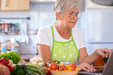 Attractive senior woman in home kitchen with vegetables on the table looking for new menu on...