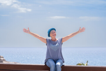 Smiling senior woman with blue cap sitting on bench close to the sea with open arms. Elderly lady relaxing enjoying vacation or retirement under the sun. Horizon over water