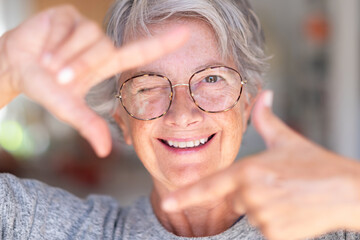 Portrait of smiling attractive senior woman with glasses gesturing the camera frame with her hands looking at the camera while winking. Funny granny makes faces