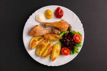 Fried chicken legs, baked potatoes, lettuce salad with red beans and cherry tomatoes, mayonnaise, mustard and ketchup on a white plate on a black background.