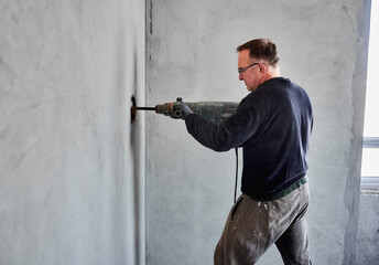 Side view of adult builder with glasses who using perforator to make hole in gray wall inside room...