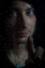 Portrait of a young hooded man pointing his finger from behind a glass covered with droplets, focus on the finger and the glass