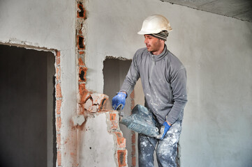 Worker in protective helmet with perforator completing deconstruction of rest of brick wall inside...