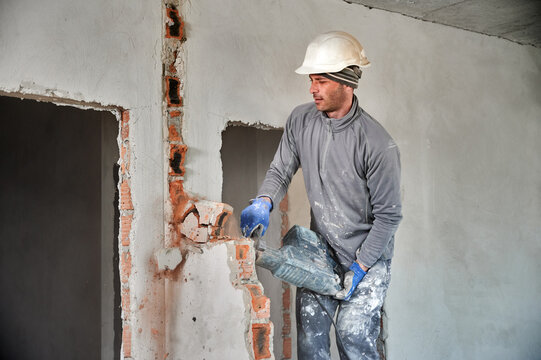 Worker in protective helmet with perforator completing deconstruction of rest of brick wall inside room. Builder carring out repair work on dismantling of walls and partitions.