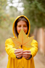 Selective focus of front close-up of unrecognizable woman holding a maple tree leaf. Vertical cropped view of woman with autumn leaf in yellow raincoat outdoors. Nature and people backgrounds.