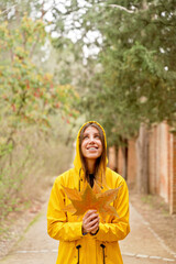 Front view of happy woman with raincoat holding maple tree leaves. Vertical mid waist view of woman looking up at fallen leaves in yellow hoodie outdoors. People and nature backgrounds.