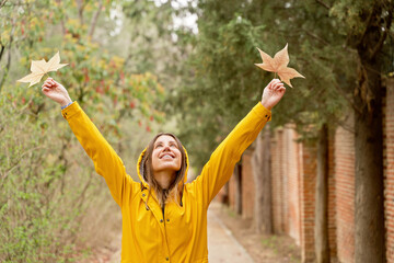 Front view of happy woman with raincoat raising arms with maple tree leaves. Horizontal mid waist view of woman looking up at fallen leaves in yellow hoodie outdoors. People and nature backgrounds.