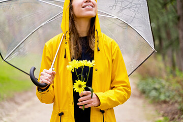 Close-up of happy woman walking with umbrella and flowers in park. Horizontal cropped view of woman in yellow raincoat and daisies bouquet on rainy park. People and nature