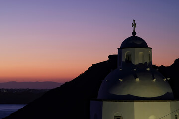 View of a beautiful whitewashed Orthodox Church and Oia in Santorini, while the sun is setting in a dramatic way - obrazy, fototapety, plakaty