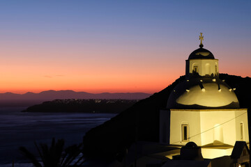 Fototapeta premium View of a beautiful whitewashed Orthodox Church and Oia Santorini in the background, while the sun is setting in a dramatic way