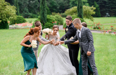 Naklejka na ściany i meble Front view of newlyweds with friends, holding wineglasses with beverage, standing together and celebrating wedding day in amazing garden
