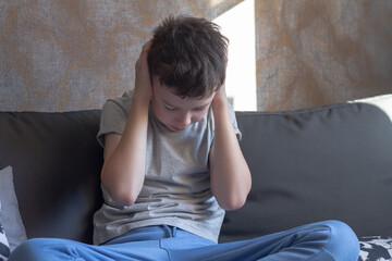 A teenage boy clutched his head with his hands, a teenage schoolboy is sitting in his room on the couch and is experiencing stress and resentment