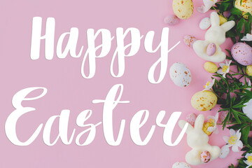 Happy Easter text and Easter chocolate eggs, bunnies and spring flowers border flat lay on pink background. Happy Easter greeting card. Seasons greeting card, handwritten lettering