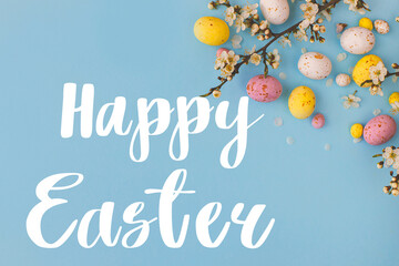 Happy Easter greeting card. Happy Easter text and colorful Easter chocolate eggs and cherry blossoms border on blue background flat lay. Seasons greeting card, handwritten lettering