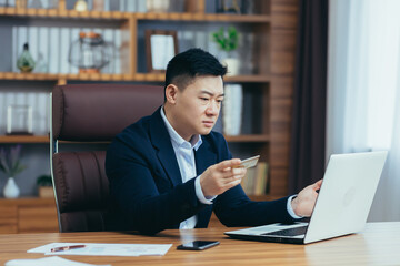 Young successful Asian man banker, banker holding a credit card, talking on a laptop on a video call, consulting, selling. looking at the camera, smiling