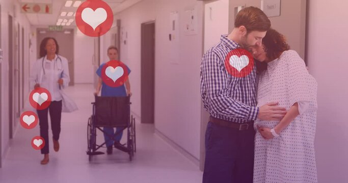 Animation of heart icons over caucasian man embracing pregnant wife in hospital