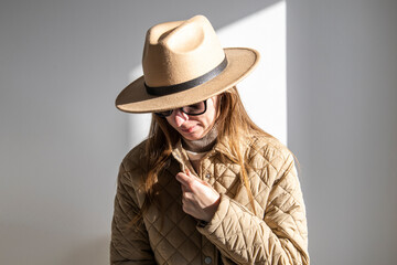 Young woman in a jacket in sunglasses in a hat against a white wall.