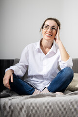 Smiling young woman in glasses in a white shirt sitting on a sofa against the background of white walls.