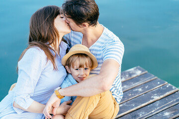 Mother and father kissing while their little son on wooden bridge over blue water on background....
