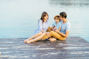 Obraz premium Portrait of middle aged father, lovely mother and their cute little son sitting on the wooden pier near the lake, at sunset on summer day.