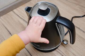 Toddler baby boy holds a hot kettle with boiling water. Child safety issues in the home room,...