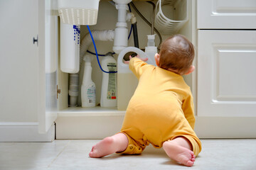 Toddler baby boy is playing with detergents and cleaning products in an open kitchen cabinet. Child...
