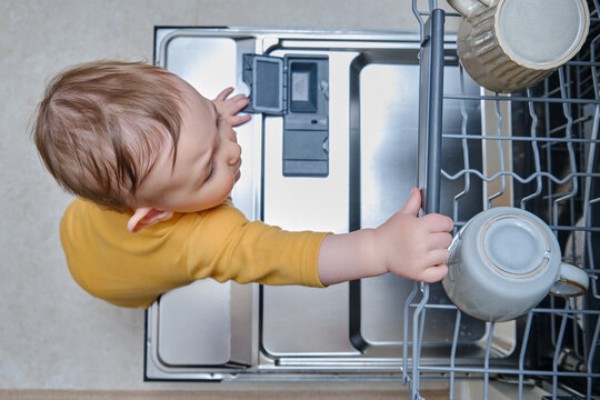 Baby Boy Toddler Is Standing By An Open Dishwasher With Dirty Dishes Inside. Child Safety Issues In The Home Room, Little Kid