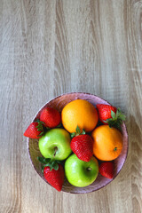 Pink bowl filled with fresh apples, oranges and strawberries on wooden table. Flat lay. 