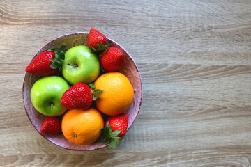 Pink bowl filled with fresh apples, oranges and strawberries on wooden table. Flat lay. 