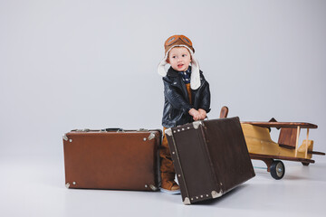 A happy boy in a helmet and a pilot's jacket carries a brown suitcase. Portrait of a child pilot, a child in a leather jacket. Little traveler with a suitcase