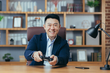 Young handsome asian man in suit in office at work playing computer games on console joystick, sitting at table, smiling, happy, relaxed